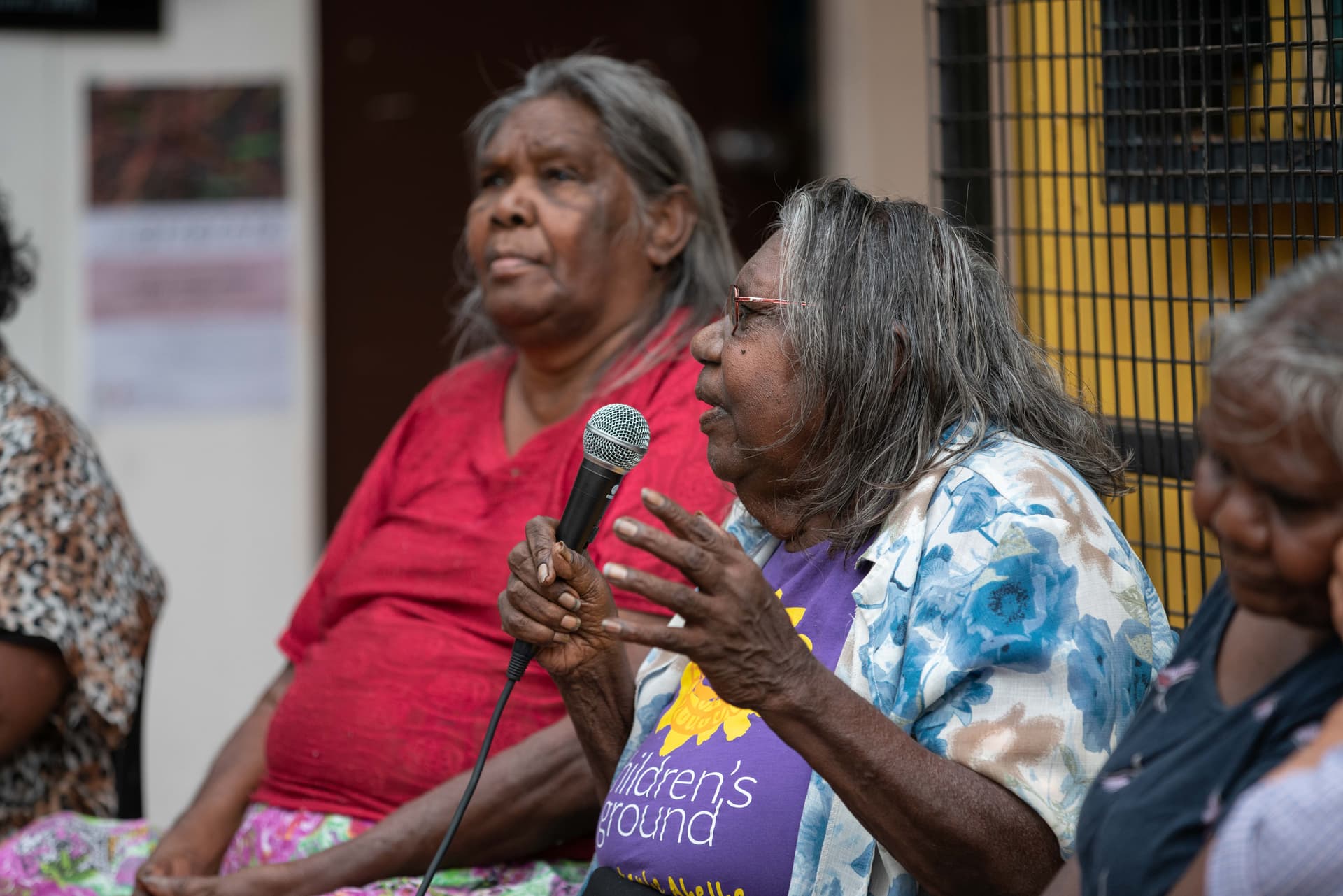 An older woman speaking into a microphone while seated among other participants.