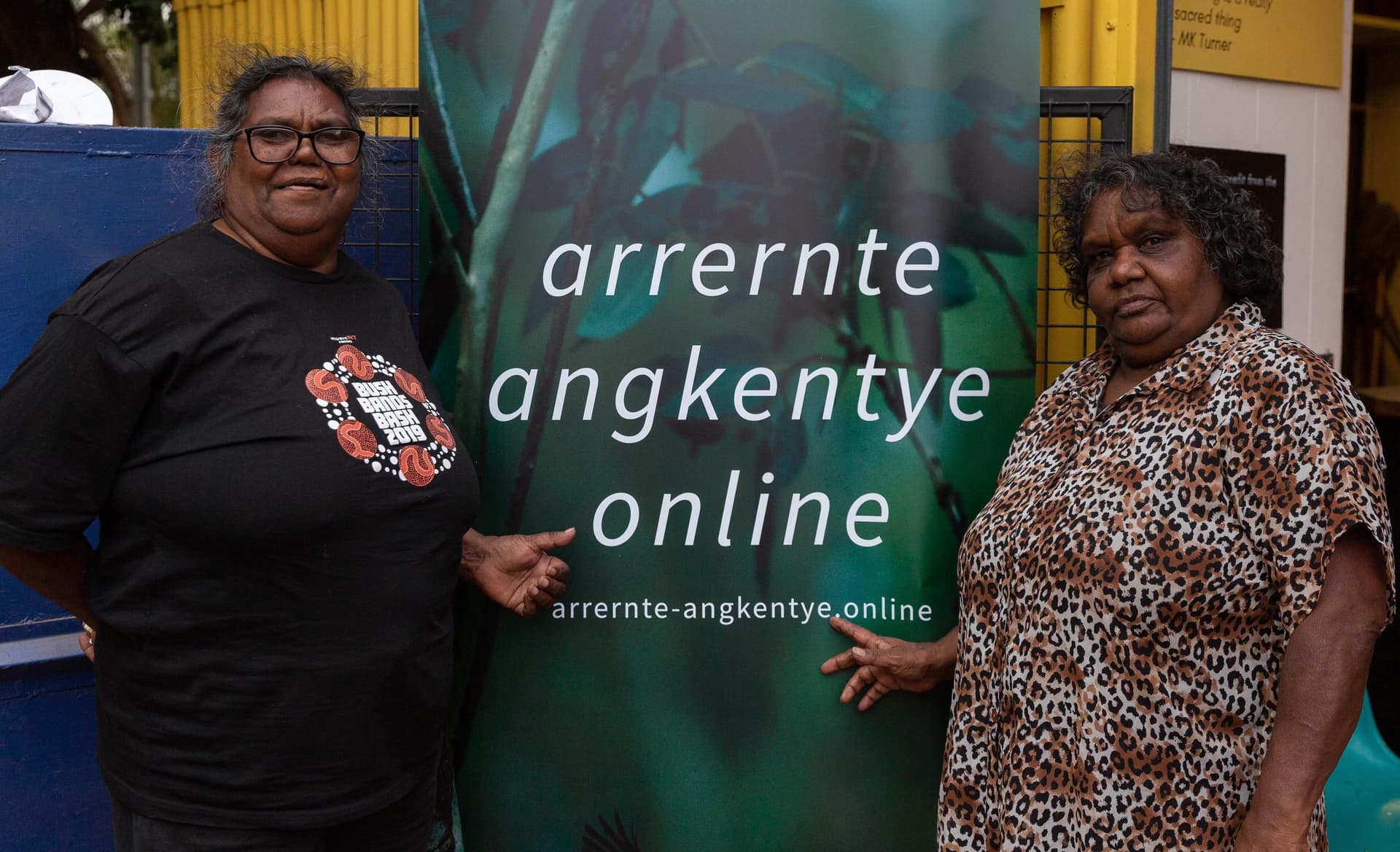 Two women standing beside a large Arrernte Angkentye Online banner.