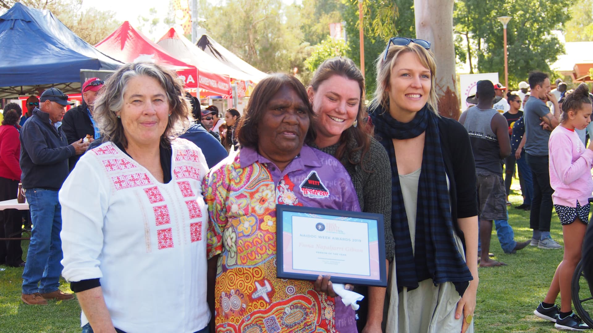 Four women standing together at an outdoor community event while holding an award certificate.